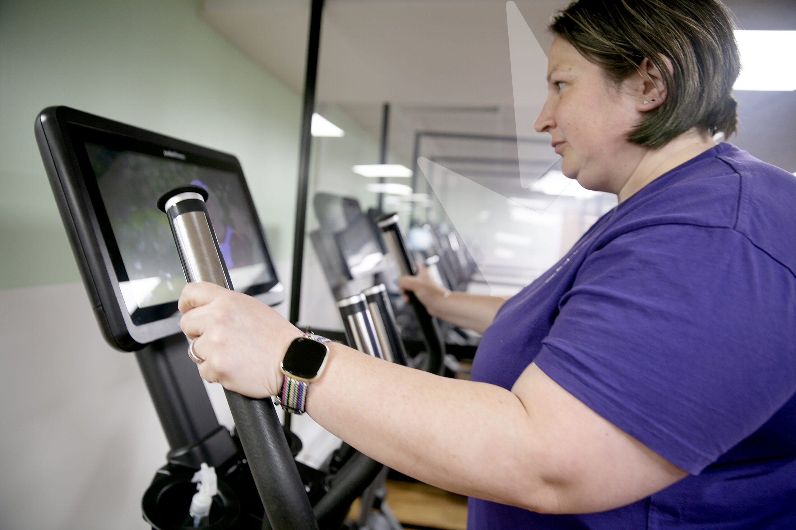 A middle aged lady wearing a purple top, watching the screen on a cross trainer